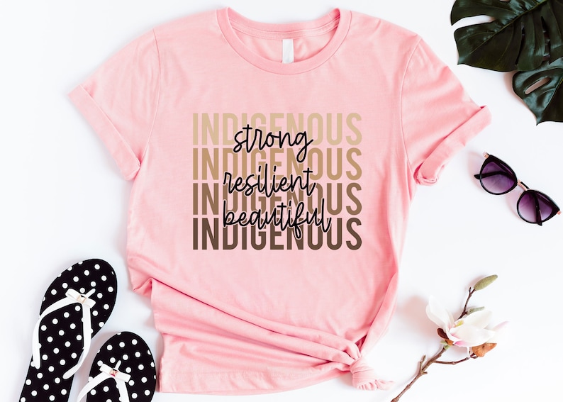 Może przedstawiać: R&oacute;żowa koszulka z nadrukiem "Indigenous strong resilient beautiful Indigenous" w kolorze brązowym i czarnym na przodzie.