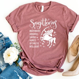 Op de afbeelding: Een mauve t-shirt met een witte afbeelding van het sterrenbeeld Boogschutter en de woorden "Boogschutter", "Onafhankelijk", "Grootmoedig", "Zorgzaam", "Eerlijk", "Optimistisch" en "Intelligent".