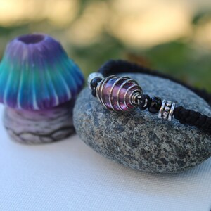 May include: A handmade bracelet with a pink glass bead encased in silver wire, black beads, and a black woven cord. A small, colorful mushroom sculpture with blue, green, and purple hues sits nearby on a stone.