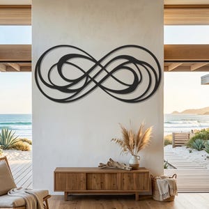 May include: A black metal wall art piece featuring an abstract, flowing infinity symbol design with multiple overlapping loops and curves, mounted horizontally on a plain white wall above a wooden console table and a vase of dried grass.