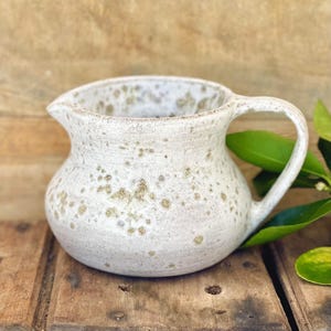 May include: A ceramic pitcher with a rounded body and a single handle. The surface is off-white with a speckled, mottled texture featuring brown and grey spots throughout. The pitcher has a small pouring spout and a wide, open top opening.