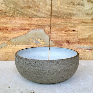 May include: A brown ceramic incense holder with a white interior. A brown incense stick is standing upright in the center of the bowl.