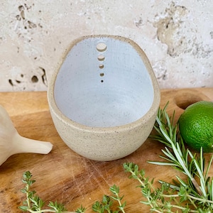 May include: A speckled beige ceramic bowl with a light blue interior. The bowl has a series of circular cutouts along the top edge. The bowl is on a wooden surface with herbs and a lime.