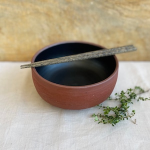 May include: A red ceramic bowl with a black interior and a rough textured exterior. The bowl is sitting on a white linen tablecloth with a pair of wooden chopsticks resting on the rim.