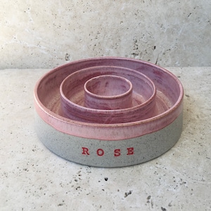 May include: A pink and grey ceramic dog bowl with three tiers. The bowl is inscribed with the word "ROSE" in red.