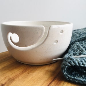May include: White ceramic yarn bowl with a speckled finish and a large opening for yarn. The bowl has a unique design with a curved opening for yarn to feed through.