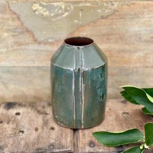 May include: A cylindrical ceramic vase with a tapered neck and a glossy, mottled dark green glaze. The surface features a vertical raised ridge running down the side, with subtle brown undertones visible along the edges and the rim of the opening.