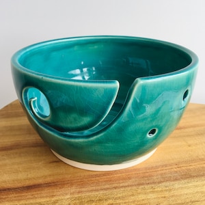 May include: A teal ceramic yarn bowl with a unique design. The bowl has a large opening for yarn and a smaller opening on the side for the yarn to feed through.