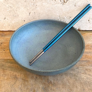 May include: A blue ceramic bowl with a matte finish, paired with a set of blue chopsticks. The chopsticks are resting inside the bowl, creating a minimalist aesthetic. The bowl is a muted blue-grey color.