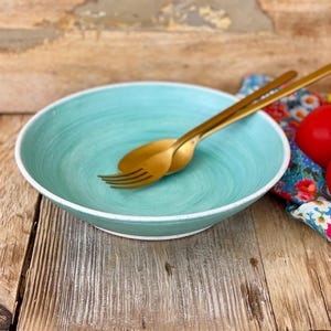 May include: A turquoise blue ceramic bowl with a white rim, sitting on a wooden surface. Two gold utensils are resting inside the bowl.