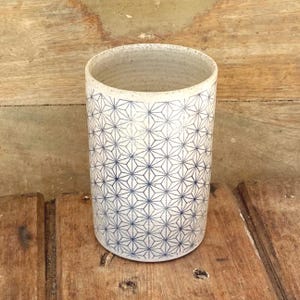 May include: A cylindrical ceramic tumbler featuring a speckled off-white base with a repeating dark blue geometric star pattern. The vessel has a smooth, straight-sided profile with a wide open top and a flat base, showcasing a clean, functional design.