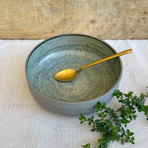 May include: A grey ceramic bowl with a green speckled glaze. A gold spoon is resting inside the bowl. The bowl is sitting on a white linen tablecloth.