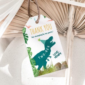 Puede incluir: Una etiqueta blanca con una cuerda marrón presenta un dinosaurio verde azulado con un gorro de fiesta. El texto dice "THANK YOU" y "TWO-REX" con el nombre "HENRY". Hojas verdes y dinosaurios voladores naranjas decoran los bordes.