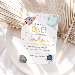 以下が含まれることがあります： カラフルなロケット、月、星が描かれた白い招待状。テキストには「5、4、3、2... ONE! ハドソンの太陽の周りを回る初めての旅を祝うために、私たちと一緒に祝ってください！ 土曜日、1月15日、午後4時 1234 Party Lane、カリフォルニア州 56789 RSVP to Mom 123 456 7890」と書いてあります。