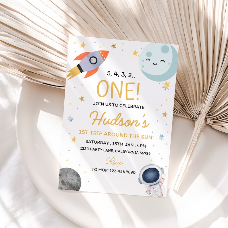 Op de afbeelding: Een witte uitnodigingskaart met een kleurrijke raket, maan en sterren. De tekst luidt "5, 4, 3, 2... EEN! Vier met ons Hudsons 1e reis rond de zon! Zaterdag 15 januari, 16:00 uur 1234 Party Lane, Californi&euml; 56789 RSVP aan Mama 123 456 7890".
