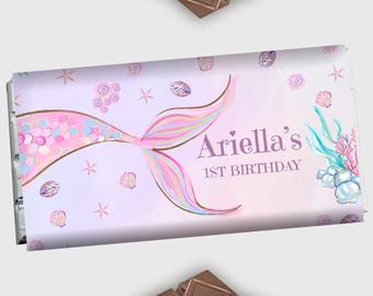 Op de afbeelding: Een paarse en roze zeemeerminstaart met gouden glitter en schelpen op een lichtpaarse achtergrond. De tekst "Ariella's 1st Birthday" is in paars geschreven.