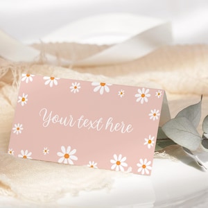 May include: A folded pink card with white daisies and orange centers. The card has the text "Your text here" in a cursive font. The card is on a white surface with a beige fabric in the background.