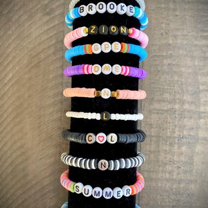 May include: A stack of colorful beaded bracelets with different names spelled out on them, including Leah, Brooke, Zion, GPE, DME, N, L, COOL, N, SUMMER, and OBX.
