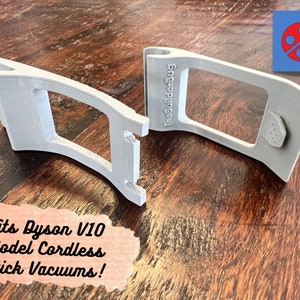 May include: Two gray 3D printed wall mounts for a Dyson V10 model cordless stick vacuum. The mounts are designed to hold the vacuum securely on a wall. The text "TheBuilder84g" is printed on one of the mounts.