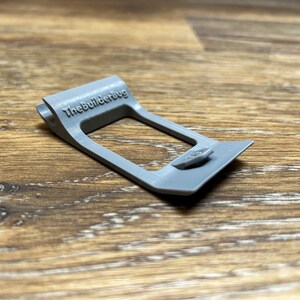 May include: A gray plastic 3D printed part with the text "TheBuilderBug" printed on it. The part has a rectangular opening and a small tab on the side.