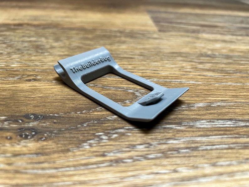 May include: A gray plastic 3D printed part with the text "TheBuilderBug" printed on it. The part has a rectangular opening and a small tab on the side.