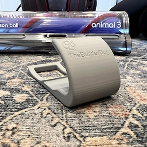 May include: A gray 3D printed stand for a Dyson Animal 3 vacuum cleaner. The stand has a cutout for the vacuum's handle and the text "The BuilderBug" printed on it.