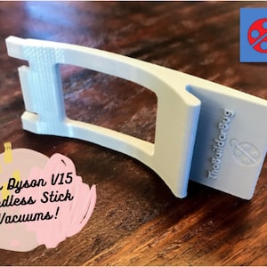 May include: White 3D printed wall mount for a Dyson V15 cordless stick vacuum. The mount has a cutout for the vacuum's handle and a logo that says "The Builder Bug". The text "Fits Dyson V15 Cordless Stick Vacuums!" is written on a pink and yellow background.