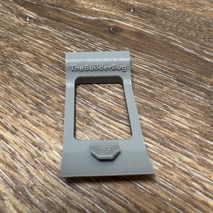 May include: A gray plastic 3D printed phone stand with the text "The Builder Bug" on the top. The stand has a small, round, gray button on the bottom.