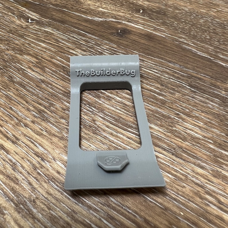 May include: A gray plastic 3D printed phone stand with the text "The Builder Bug" on the top. The stand has a small, round, gray button on the bottom.