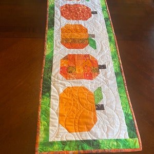 May include: A white table runner with a green and orange border features five orange pumpkins with green stems. The pumpkins are quilted with a swirl pattern.
