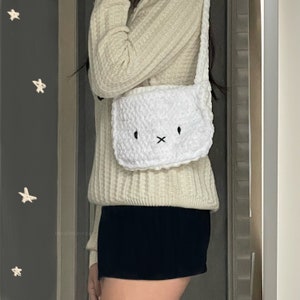 May include: White crocheted shoulder bag with a cute bunny face design. The bag has a long strap and is worn over the shoulder.