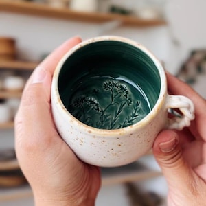 Puede incluir: Una taza de cerámica blanca con un esmalte moteado y un interior verde. El interior de la taza presenta un diseño estampado de una rama de flores verdes.