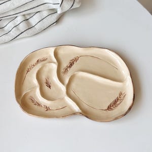Può includere: Un piatto da portata in ceramica beige con tre sezioni e disegni di spighe di grano. Il piatto è su una superficie bianca con un panno a righe sullo sfondo. Le spighe di grano sono marroni.