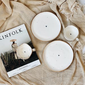May include: A still-life image featuring white ceramic plates and a mug with a small black heart design. The plates have a brown rim. A magazine with the word "KINFOLK" is also in the image. The background is a beige fabric.