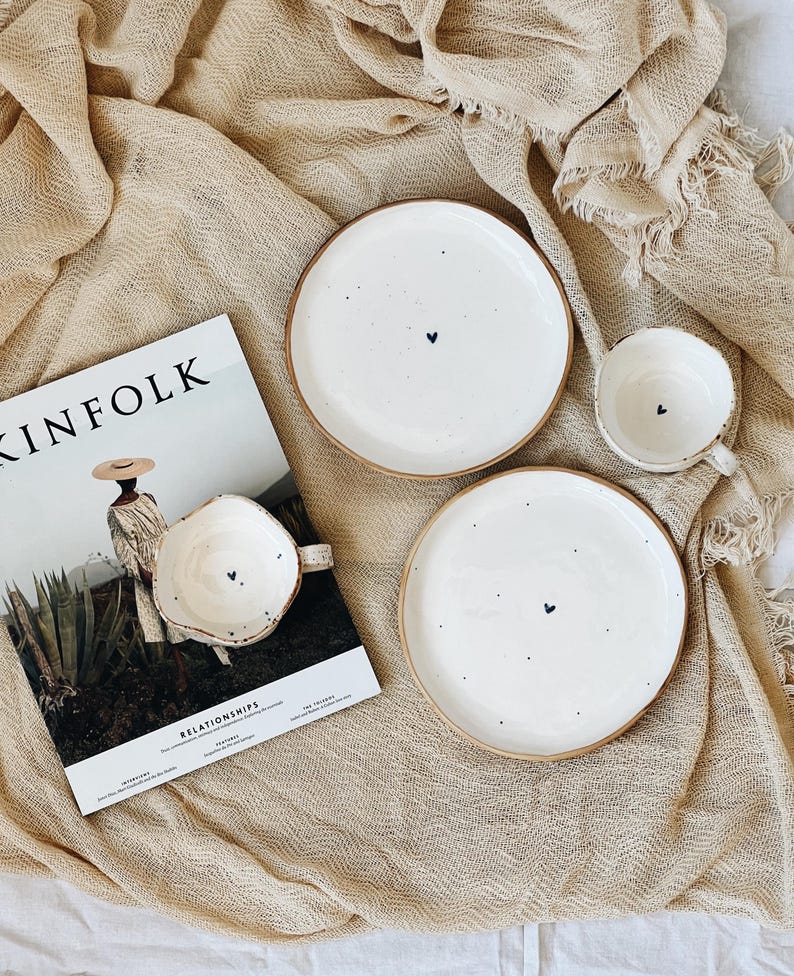 May include: A still-life image featuring white ceramic plates and a mug with a small black heart design. The plates have a brown rim. A magazine with the word "KINFOLK" is also in the image. The background is a beige fabric.