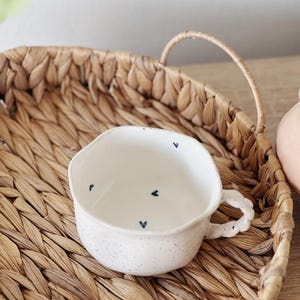 May include: A white ceramic cup with a speckled glaze and a small handle. The cup has a blue heart design on the inside.