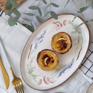 May include: An oval ceramic serving platter with floral designs, holding two custard tarts with orange slices. The platter has a brown rim and is decorated with lavender, pink, and green botanical illustrations. A gold fork and knife are on the left.