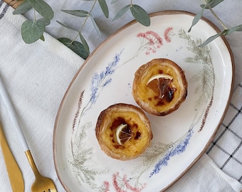 May include: An oval ceramic serving platter with floral designs, holding two custard tarts with orange slices. The platter has a brown rim and is decorated with lavender, pink, and green botanical illustrations. A gold fork and knife are on the left.