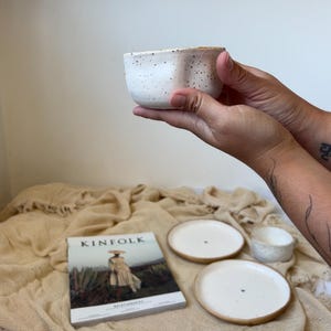 May include: A white ceramic bowl with black speckles is held in front of a soft, beige background. Two white plates with a brown rim and a Kinfolk magazine are also visible, suggesting a home decor or dining theme.