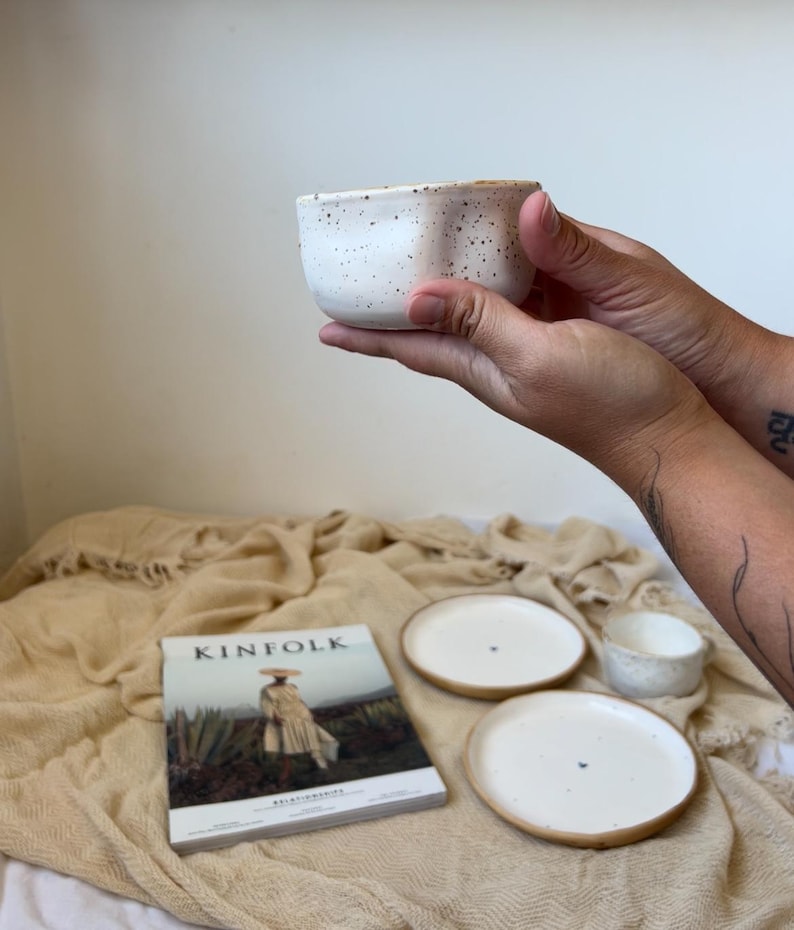 May include: A white ceramic bowl with black speckles is held in front of a soft, beige background. Two white plates with a brown rim and a Kinfolk magazine are also visible, suggesting a home decor or dining theme.
