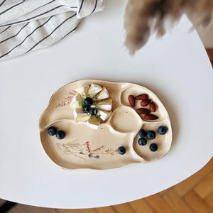 Puede incluir: Una bandeja de cerámica beige con varios compartimentos, que contiene queso brie, arándanos y almendras. La bandeja tiene un diseño floral y está colocada sobre una mesa blanca. Un paño a rayas está en el fondo.