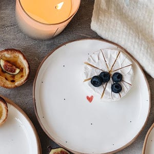 以下が含まれることがあります： 黒い斑点と赤いハートのデザインが施された白いセラミックプレート。プレートには、ブルーベリーをトッピングした丸いブリーチーズがあります。