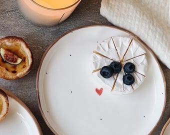May include: A white ceramic plate with black speckles and a red heart design. The plate has a round brie cheese wheel with blueberries on top.