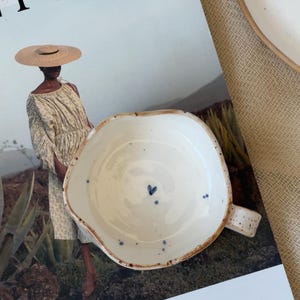 May include: A handmade, off-white ceramic bowl with a small handle and irregular edges. The bowl features a blue heart and blue speckles inside. The bowl is sitting on a magazine with the word "FOLK" in large black letters.