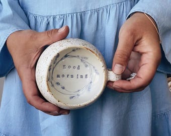 May include: A white ceramic mug with a speckled glaze and a floral design. The mug has a handle and says "Good morning" in black lettering.