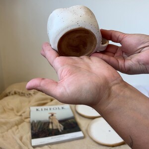 May include: A speckled white ceramic mug with a rounded shape and a small handle. The interior and base are a warm brown color. The mug is held in two hands, showcasing its design and form.