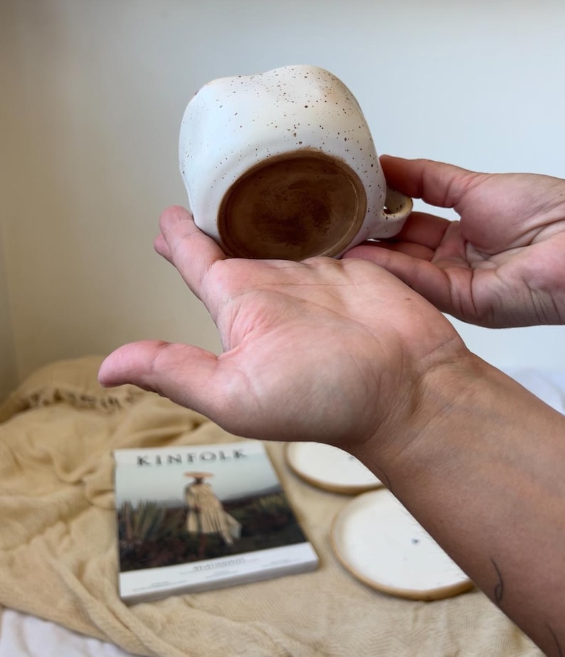 May include: A speckled white ceramic mug with a rounded shape and a small handle. The interior and base are a warm brown color. The mug is held in two hands, showcasing its design and form.