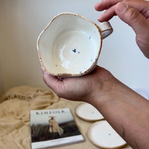 May include: Handmade ceramic bowl with an irregular rim, speckled exterior, and a small blue heart detail inside. The bowl is held in a person's hands. The interior is white, and the exterior has a natural, speckled finish.
