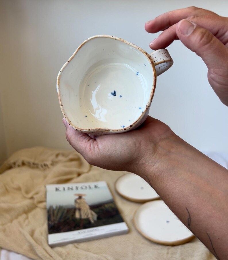 May include: Handmade ceramic bowl with an irregular rim, speckled exterior, and a small blue heart detail inside. The bowl is held in a person's hands. The interior is white, and the exterior has a natural, speckled finish.
