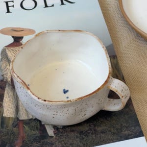May include: A speckled white ceramic mug with a brown rim and handle. The mug has a unique, irregular shape and a small blue heart detail. The mug is placed on a magazine with the word "FOLK" visible.